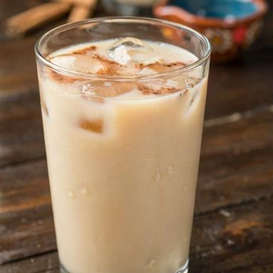 A tall glass of Mexican horchata over ice on a dark wooden table, the pale ivory rice drink dusted with ground cinnamon, with a pitcher of it and loose canela sticks behind.