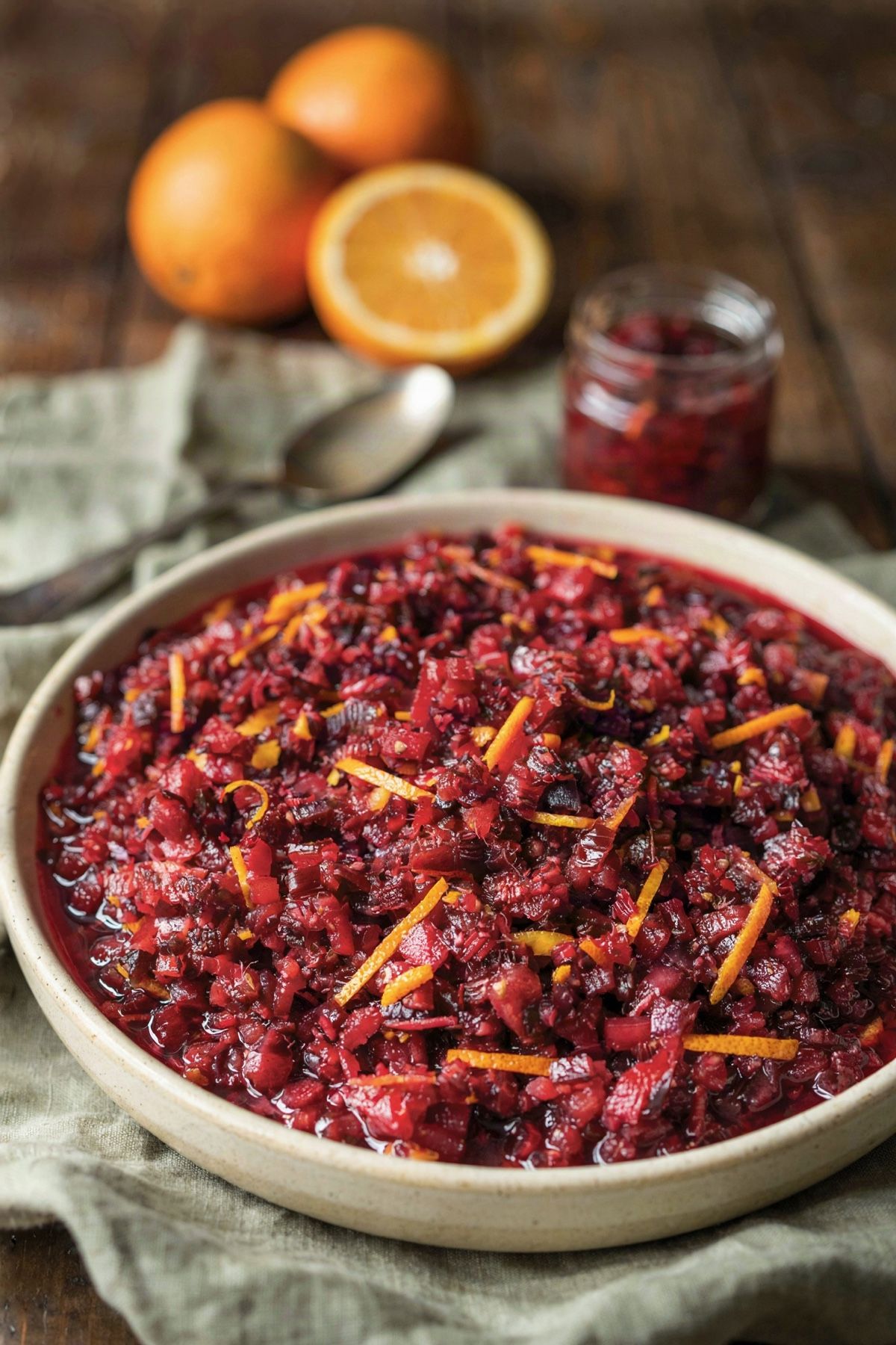 Cranberry-orange relish in a cream stoneware bowl, raw cranberries and a whole orange ground coarse and shot through with bright shreds of orange peel, with fresh oranges and a jar of relish on the table behind