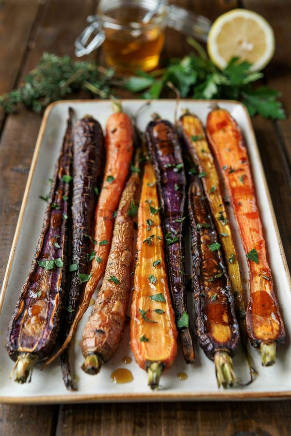 Roasted rainbow carrots on a white platter, purple, orange and gold halves caramelized and glazed with honey, scattered with chopped thyme and parsley