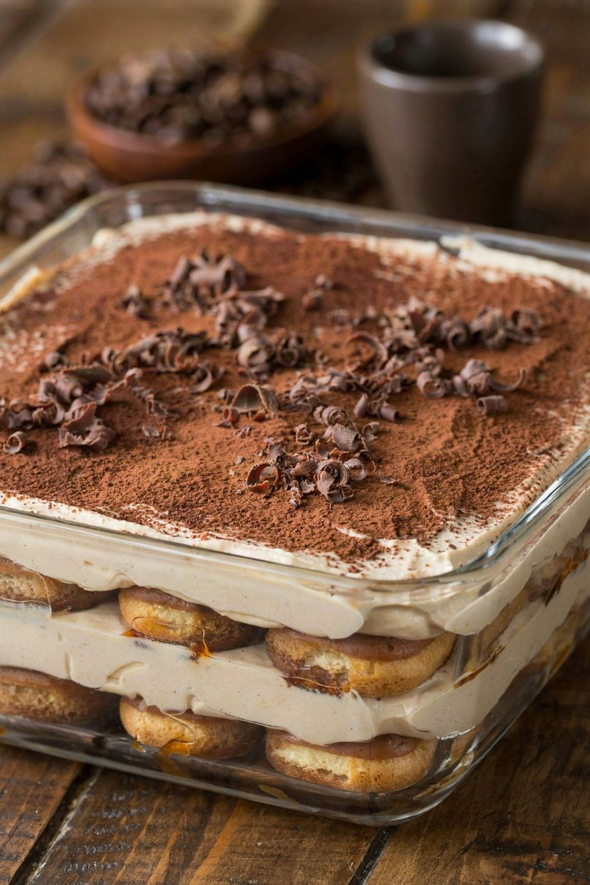 Vegan tiramisu in a square glass dish, coffee-soaked ladyfingers layered with pale cashew and coconut cream, dusted with cocoa and scattered with dark chocolate curls.