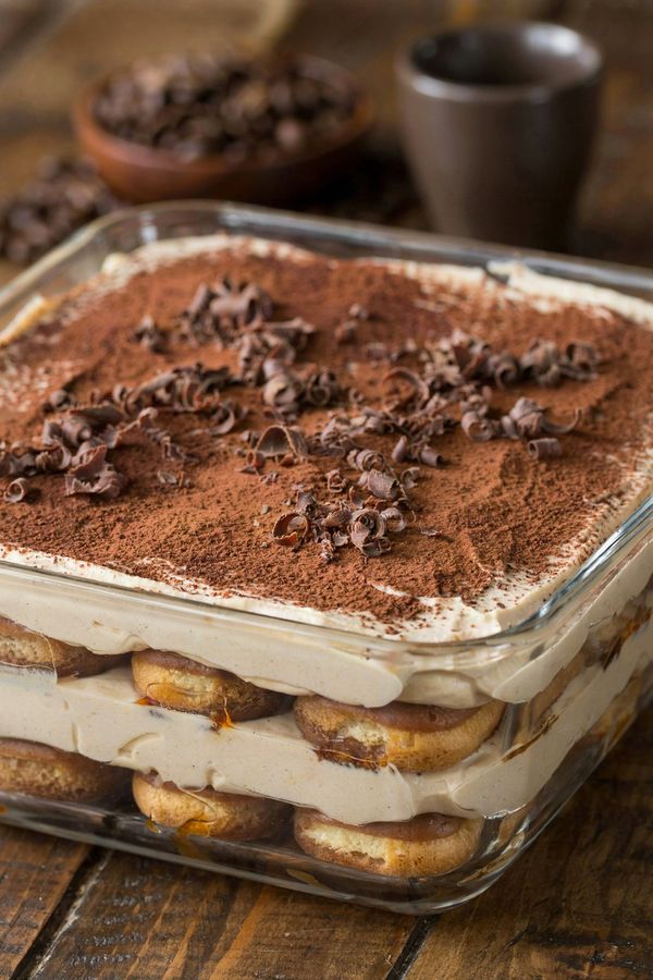 Vegan tiramisu in a square glass dish, coffee-soaked ladyfingers layered with pale cashew and coconut cream, dusted with cocoa and scattered with dark chocolate curls.