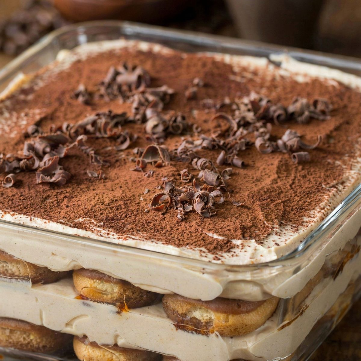 Vegan tiramisu in a square glass dish, coffee-soaked ladyfingers layered with pale cashew and coconut cream, dusted with cocoa and scattered with dark chocolate curls.