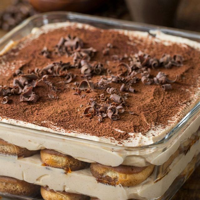 Vegan tiramisu in a square glass dish, coffee-soaked ladyfingers layered with pale cashew and coconut cream, dusted with cocoa and scattered with dark chocolate curls.