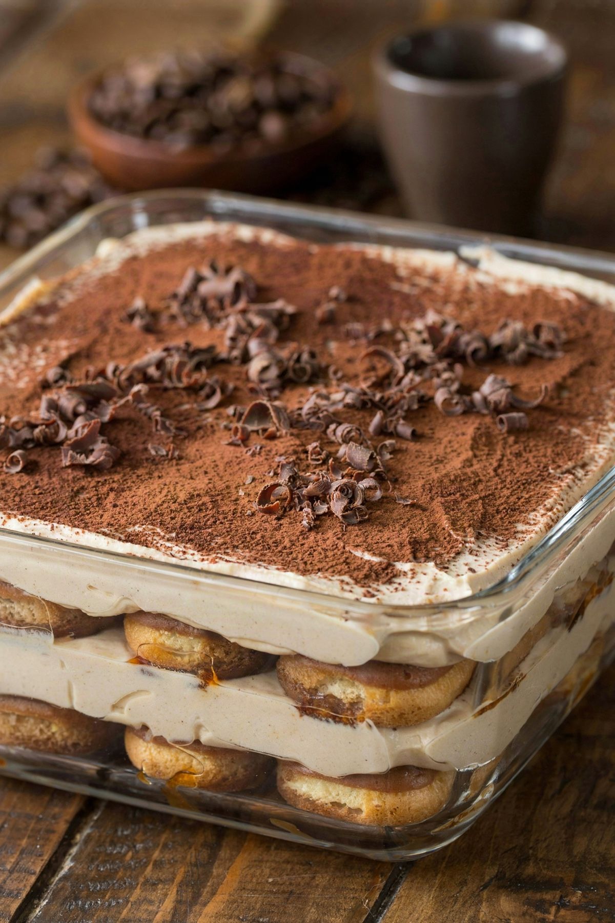 Vegan tiramisu in a square glass dish, coffee-soaked ladyfingers layered with pale cashew and coconut cream, dusted with cocoa and scattered with dark chocolate curls.