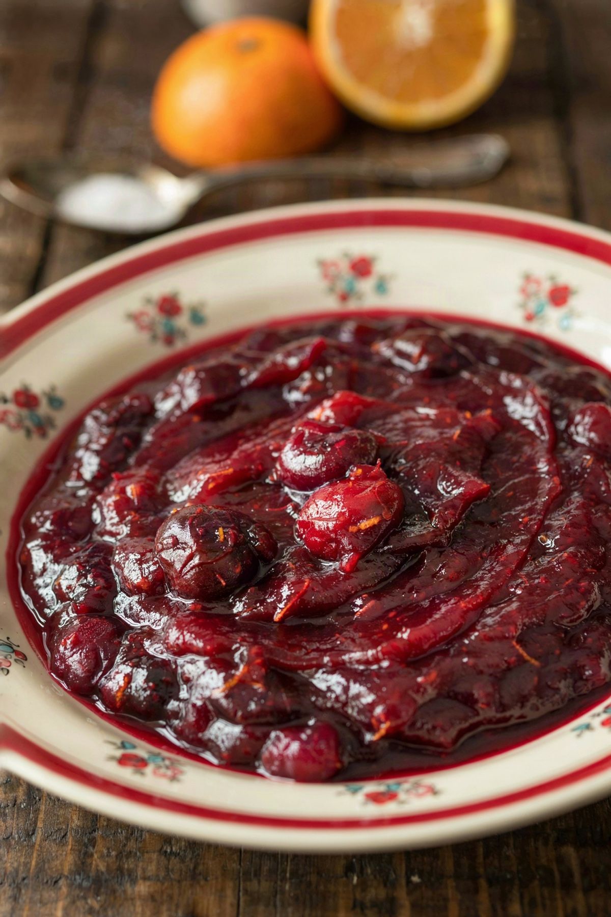 Cranberry orange sauce set thick in a cream floral bowl, dark crimson and glossy with burst berries and flecks of orange zest