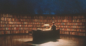 A person sits at a desk with a lamp, surrounded by tall bookshelves and a starry ceiling.