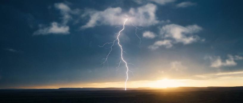 Lightning striking the ground under stormy clouds with a sunset on the horizon.