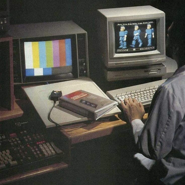 A person works at a desk with two vintage computer monitors, one displaying color bars and the other early 3D blocky figures, with a "CYBERPUNK" book nearby.
