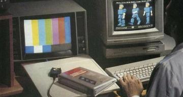 A person works at a desk with two vintage computer monitors, one displaying color bars and the other early 3D blocky figures, with a "CYBERPUNK" book nearby.
