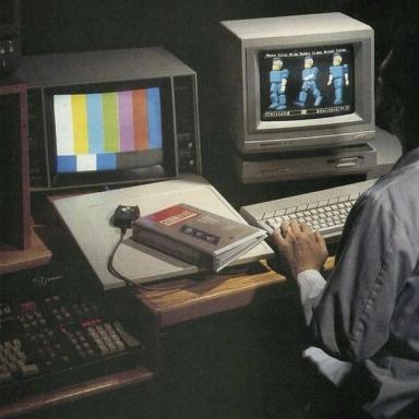 A person works at a desk with two vintage computer monitors, one displaying color bars and the other early 3D blocky figures, with a "CYBERPUNK" book nearby.