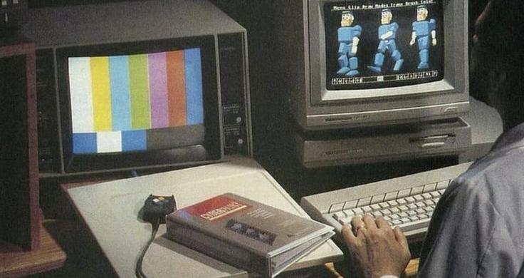 A person works at a desk with two vintage computer monitors, one displaying color bars and the other early 3D blocky figures, with a "CYBERPUNK" book nearby.