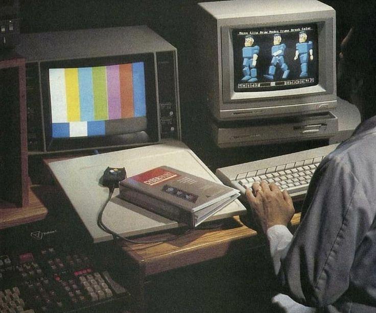 A person works at a desk with two vintage computer monitors, one displaying color bars and the other early 3D blocky figures, with a "CYBERPUNK" book nearby.