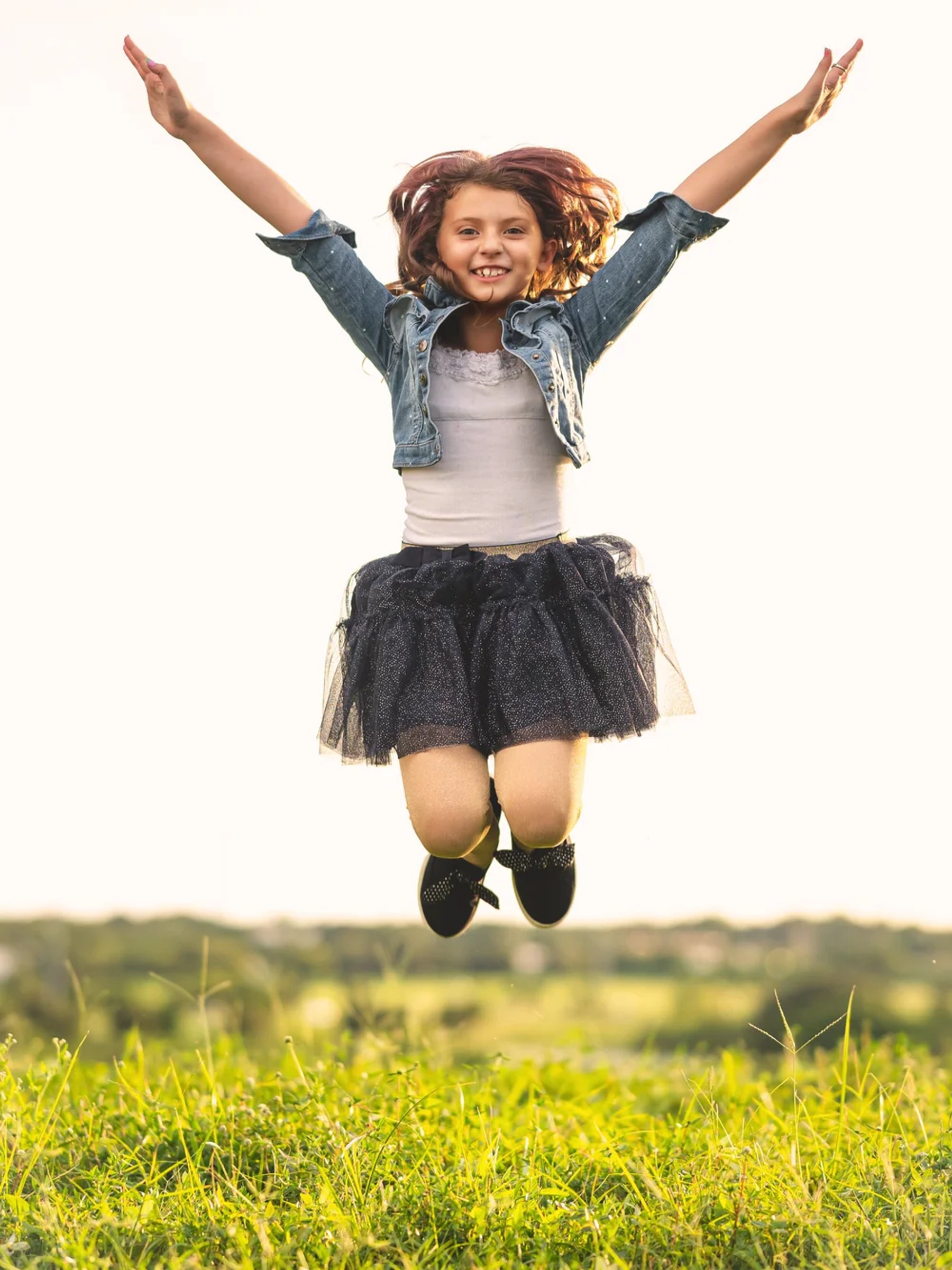 Smiling girl in a denim jacket and dark skirt jumping above the grass with both arms raised.