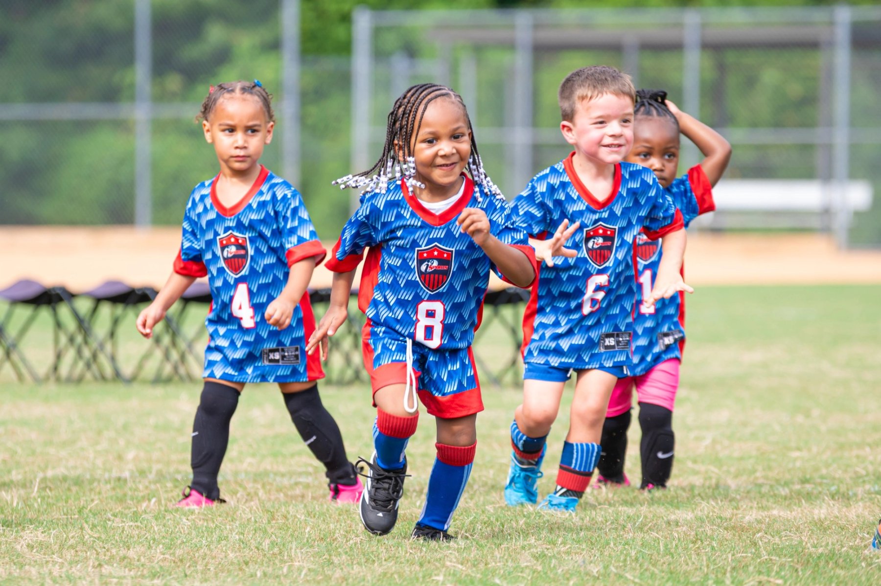 Four children wearing blue and red numbered sports uniforms move across a grassy field, with a chain-link fence and folding seats behind them.