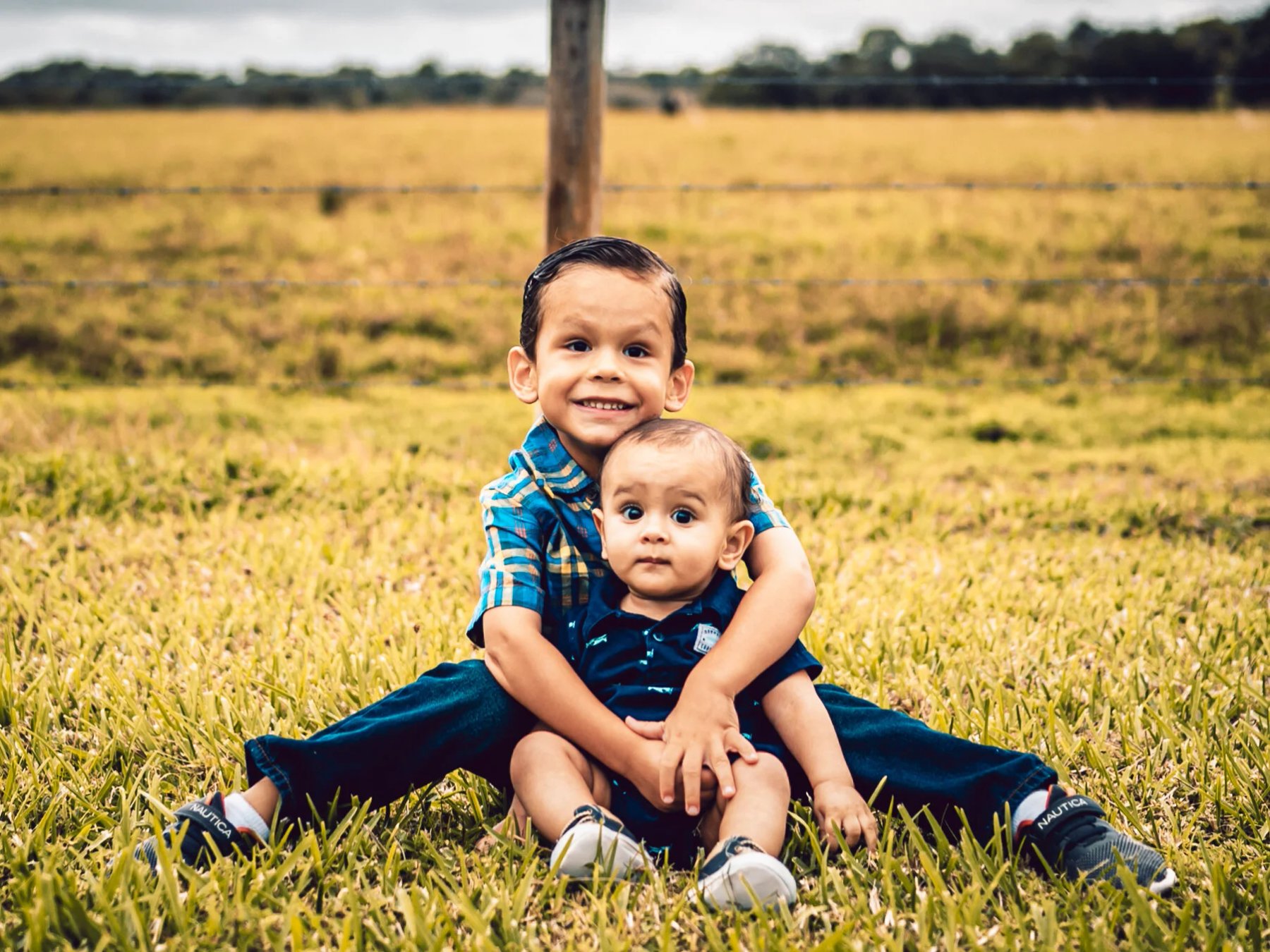 Smiling child holding a baby while seated in the grass, with a wire fence and open field behind them.