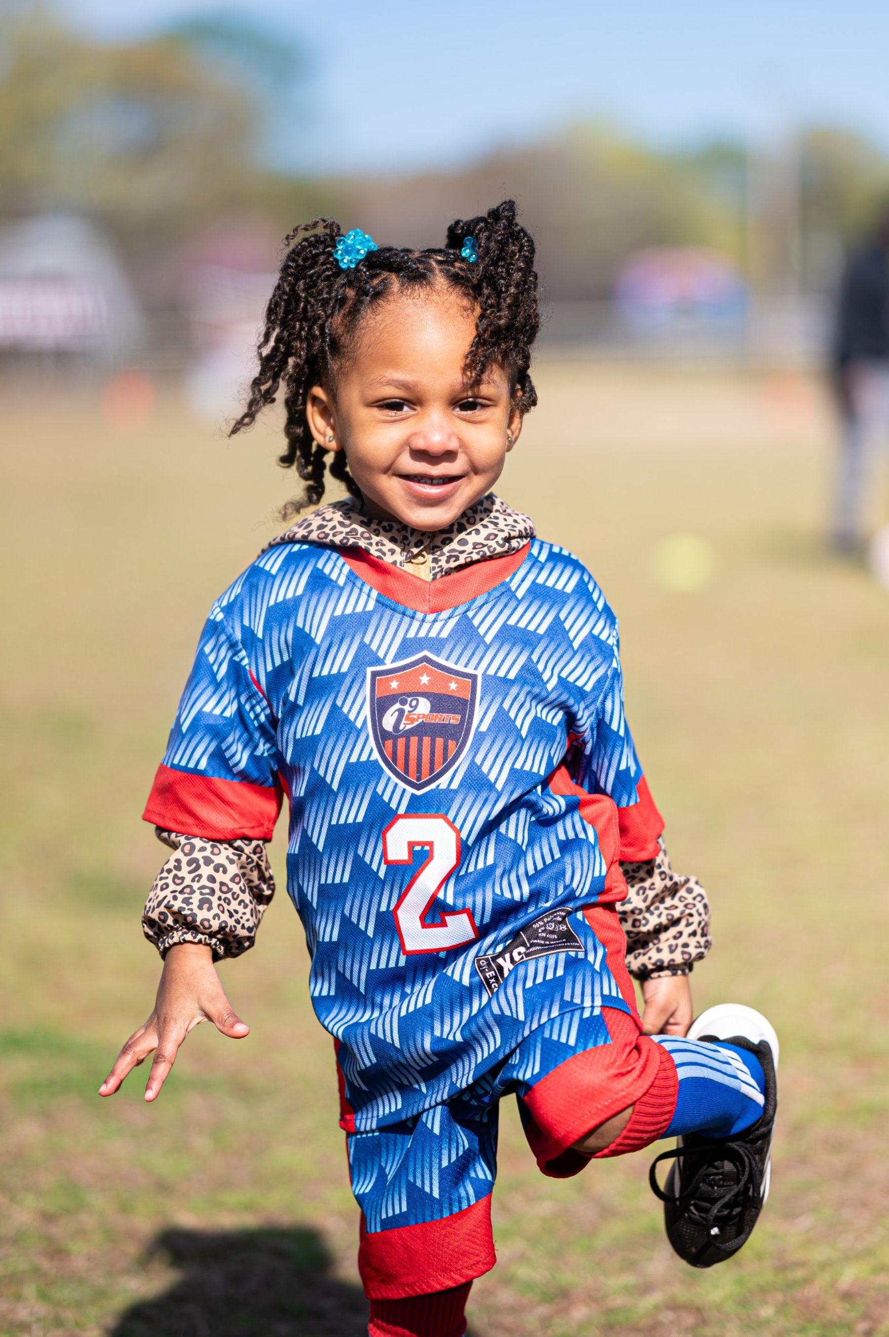 A smiling child wearing a blue and red number 2 sports uniform lifts one foot behind them on a grassy field.