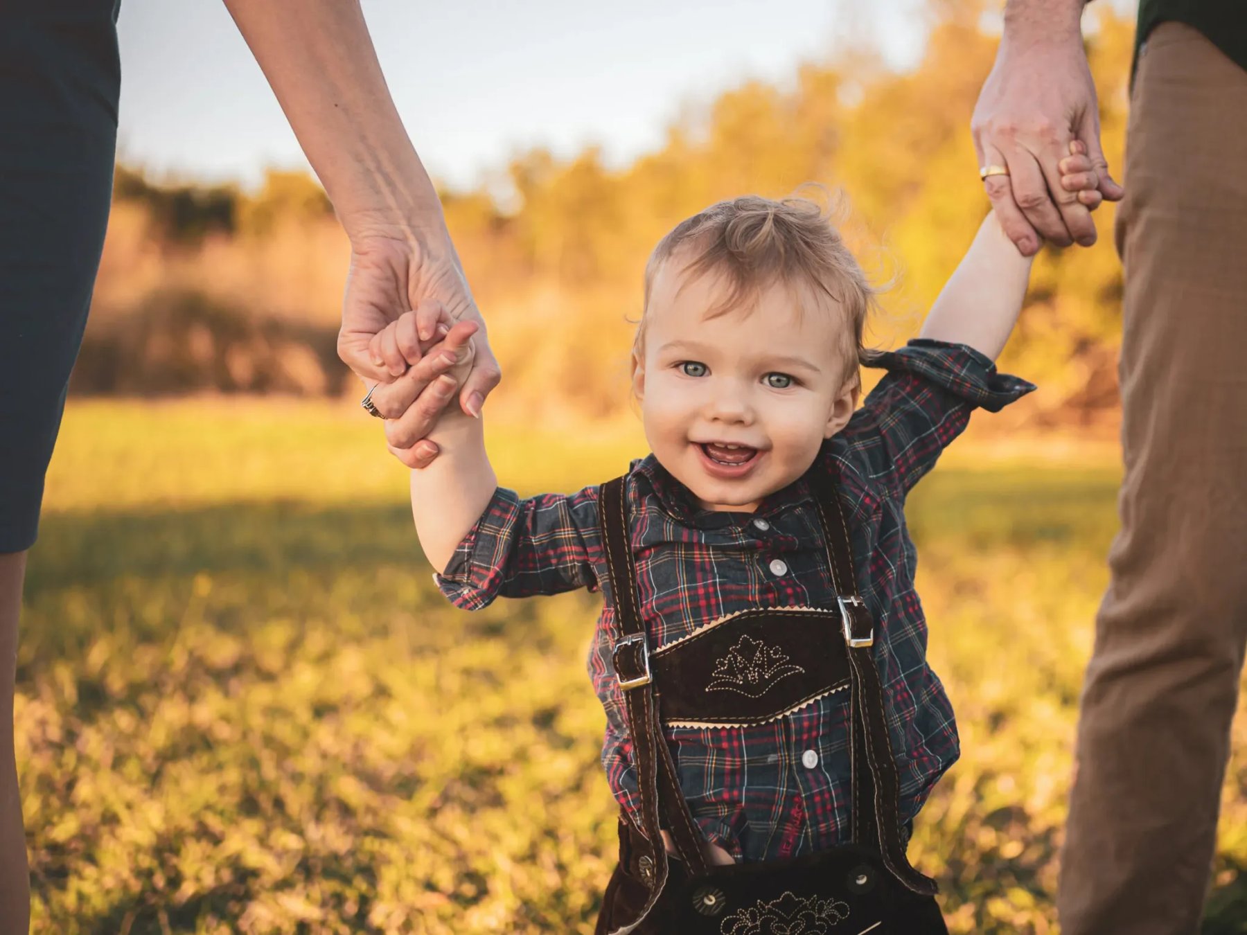 A smiling child in a plaid shirt and dark overalls holds the hands of two adults while standing in a sunlit grassy field.