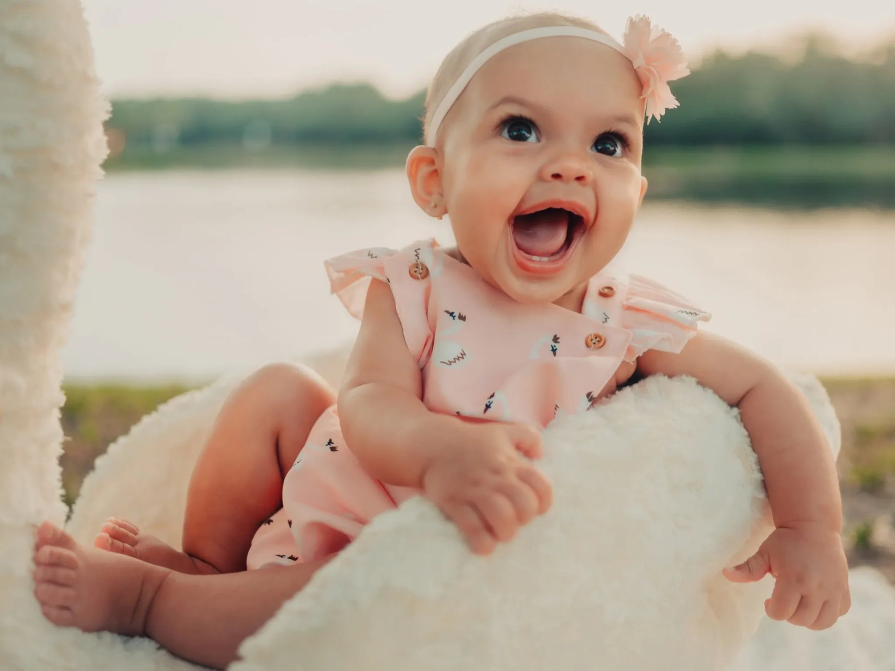 A baby with an open-mouthed smile wears a pale pink outfit and flower headband while sitting in a white plush swan beside the water.