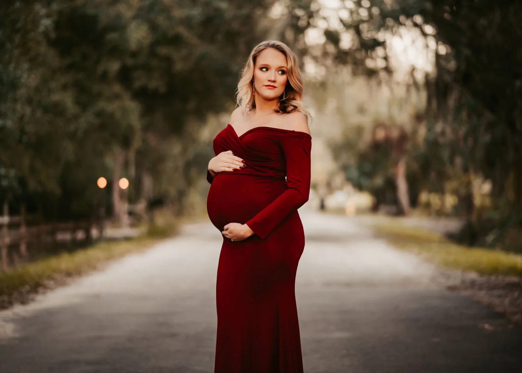 An expectant mother in a burgundy gown on a tree-lined road