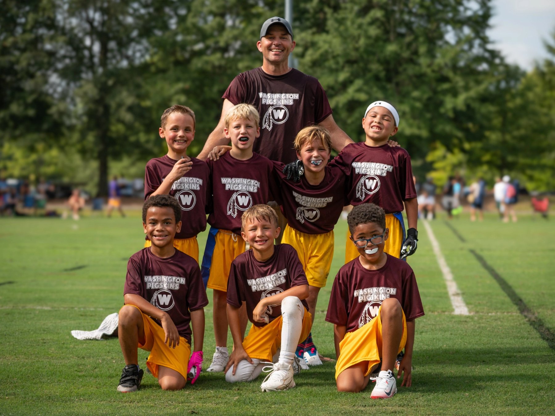 Seven children in burgundy shirts and yellow shorts pose on a marked grass field with an adult standing behind them in a matching shirt.