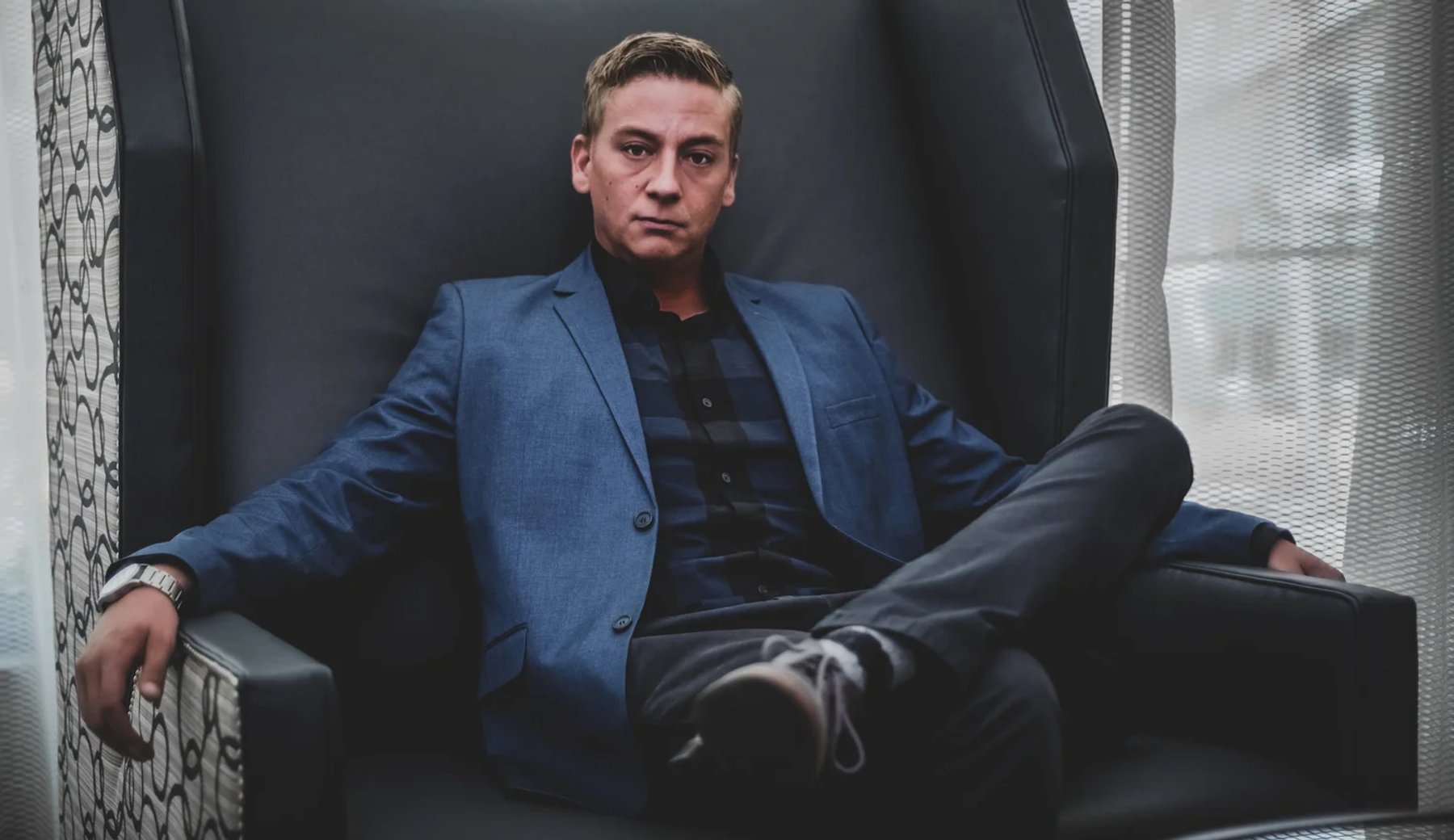 An individual portrait of a man seated beside a window in a blue suit