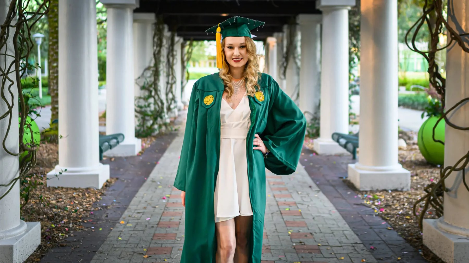 A smiling graduate in a green cap and gown with a gold tassel stands along a brick walkway framed by white columns and climbing vines.