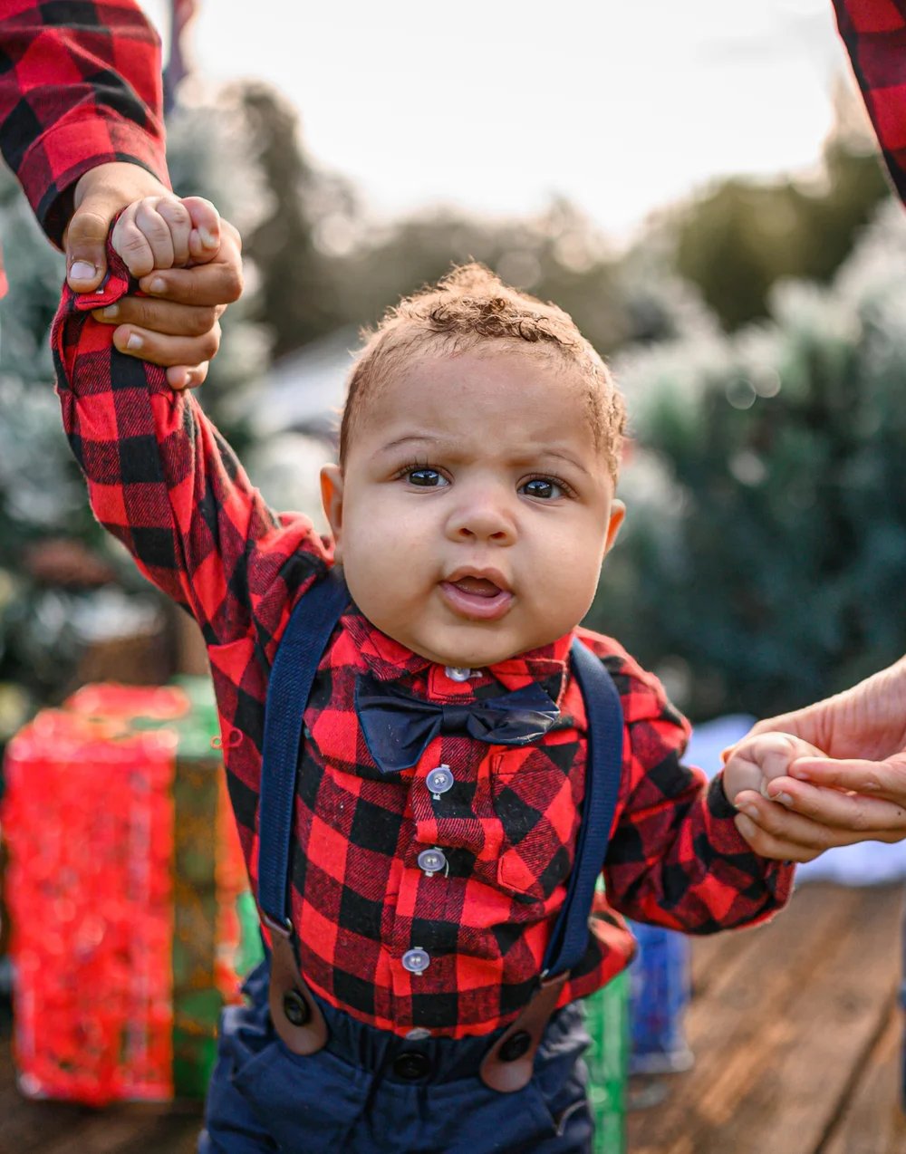 Young child in a red plaid shirt, navy bow tie and suspenders holding adults' hands in front of holiday decorations.