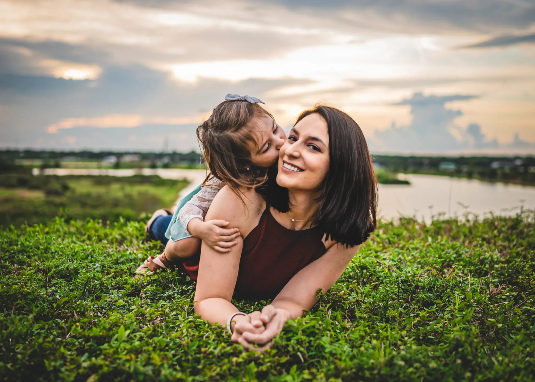 Young girl kissing a smiling woman on the cheek as they lie on grass overlooking the water.