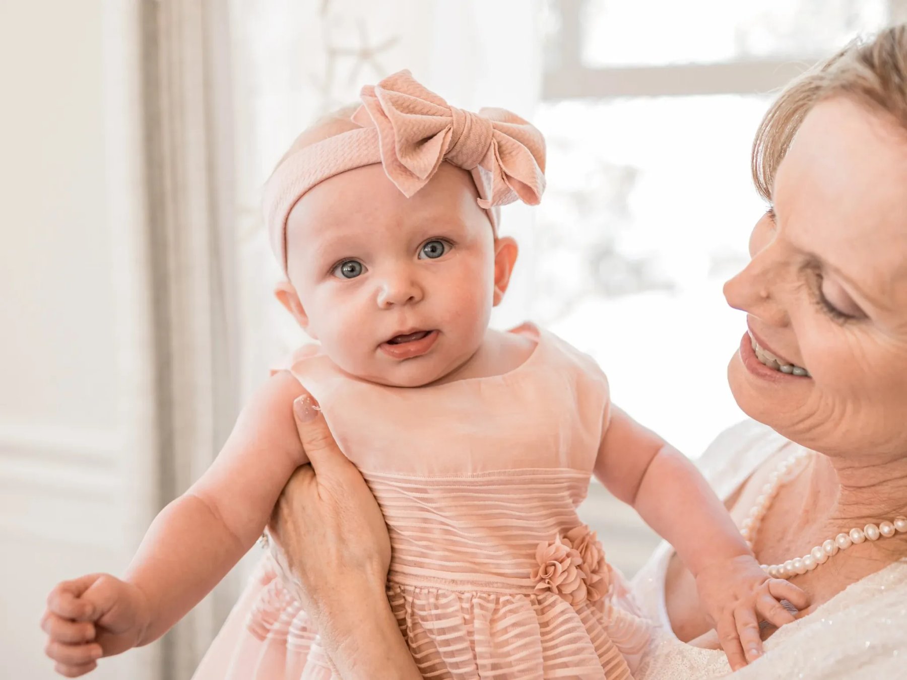 Baby in a pale pink dress and bow held beside a bright window by a smiling woman.