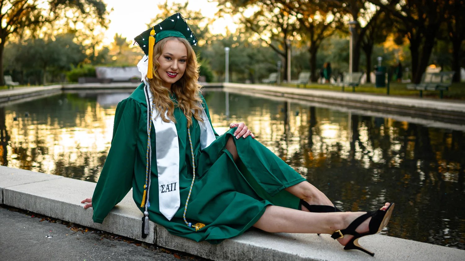 A graduate in a green cap and gown beside a pond