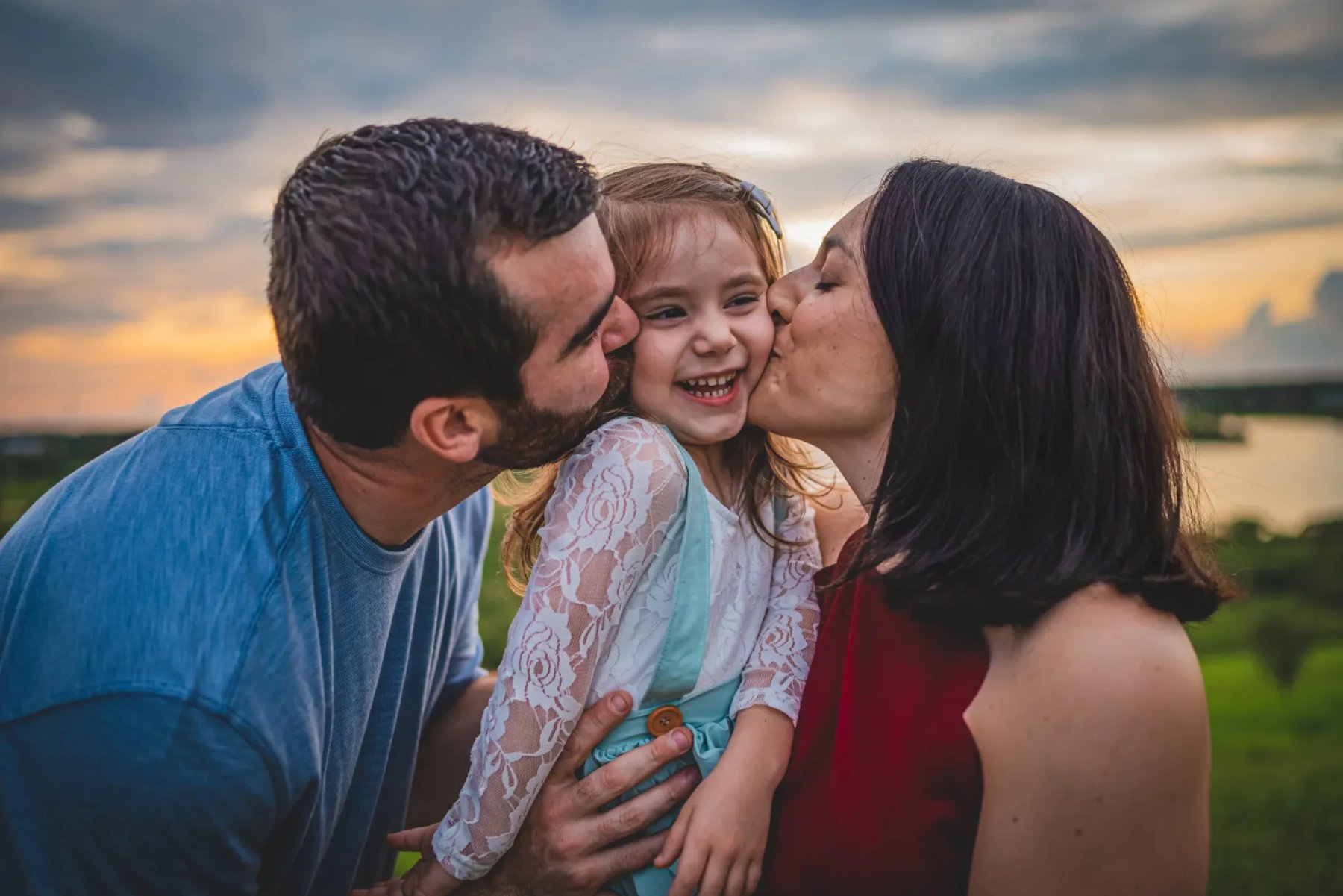 Two adults kissing a laughing child's cheeks outdoors beneath a golden, cloudy sky.
