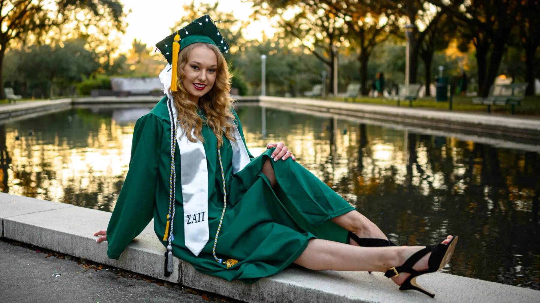 Graduate in a green cap and gown seated on a stone edge beside a reflecting pool.