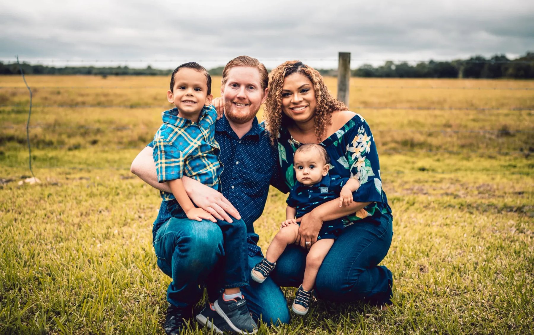 Two adults holding two children, wearing blue clothing and posing together on grass beside a wire fence.