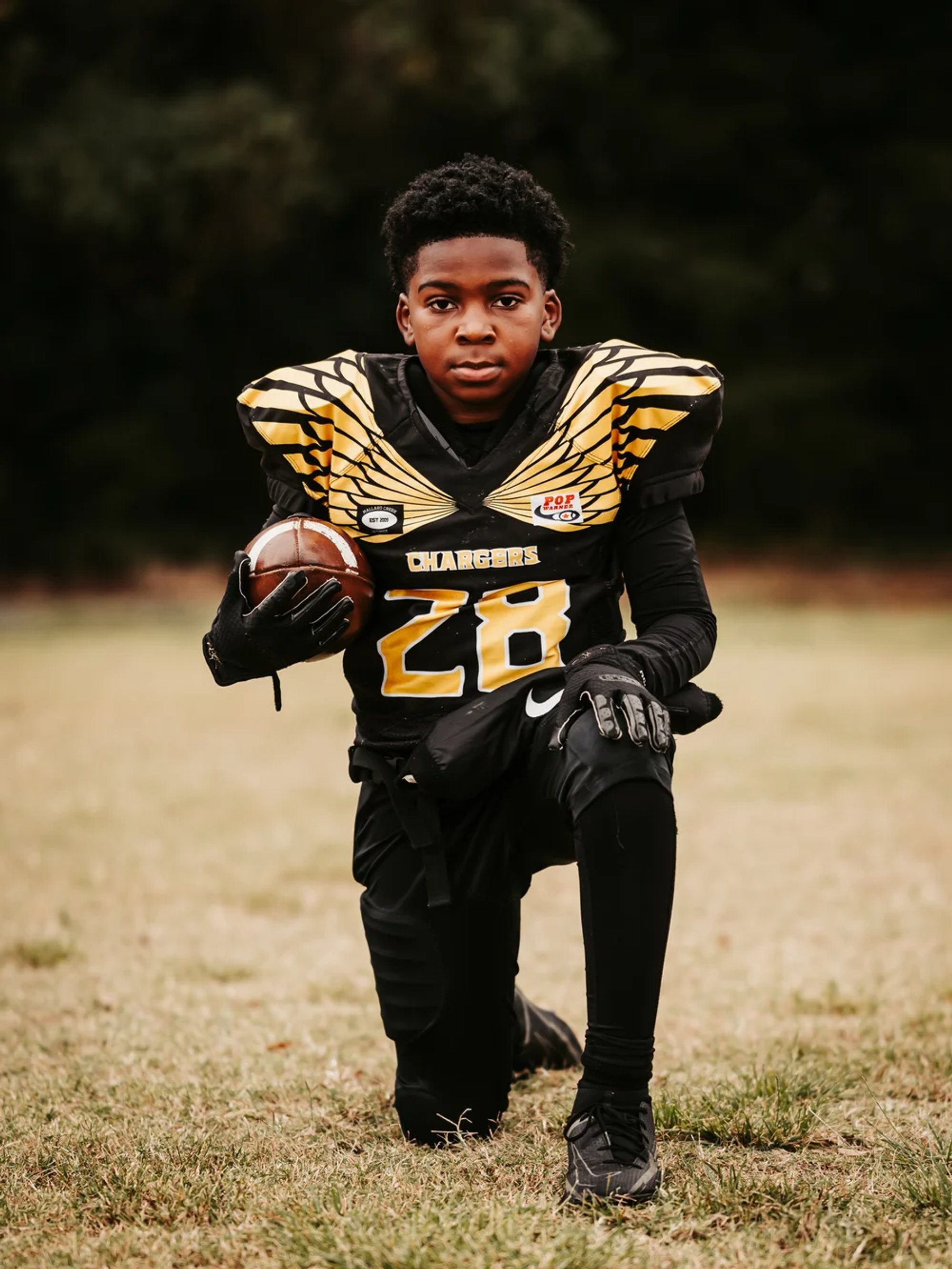 A young athlete in a black-and-gold number 28 football uniform kneels on one knee, holding a football against their side on a grassy field.