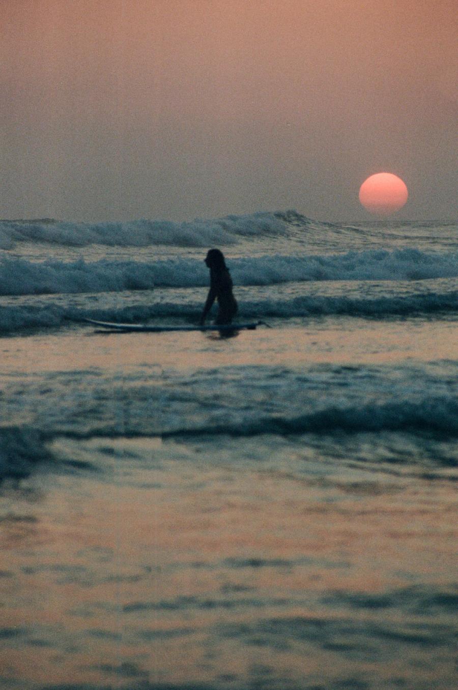 A telephoto shot of the pink red sun setting with a woman in foreground walking amongst the waves.