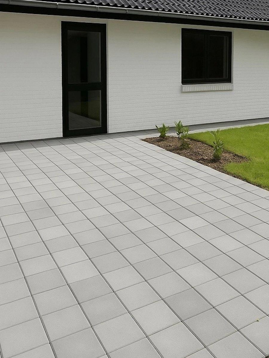 Terrasse i lyse fliser langs hvidt hus med bede og plæne i haven