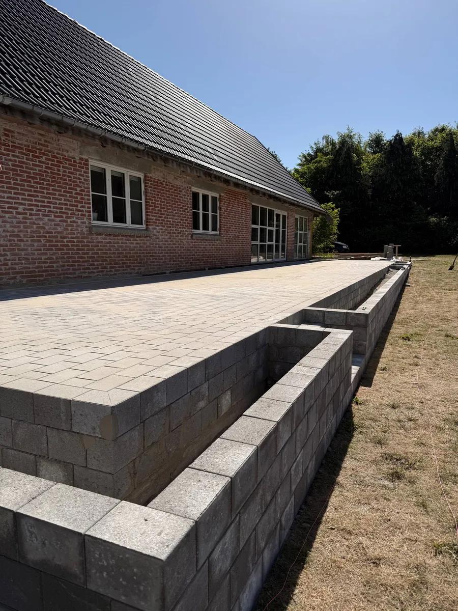 Flisebelagt terrasse langs murstenshus med støttemur af betonblokke
