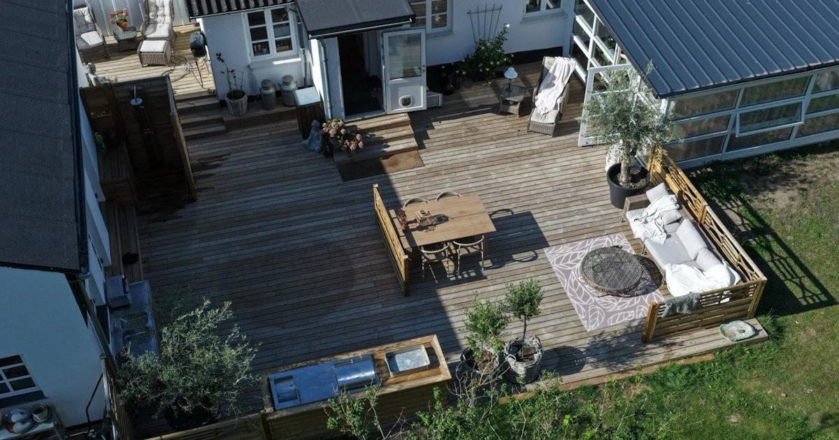 Stor terrasse i træ set fra oven med havemøbler og oliventræer i krukker