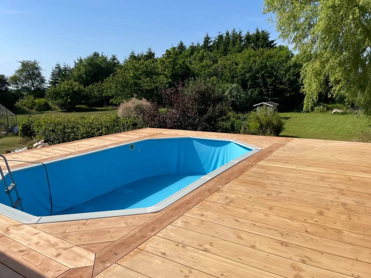 Træterrasse omkring pool i haven med lærketræsplanker og grønne træer i baggrunden