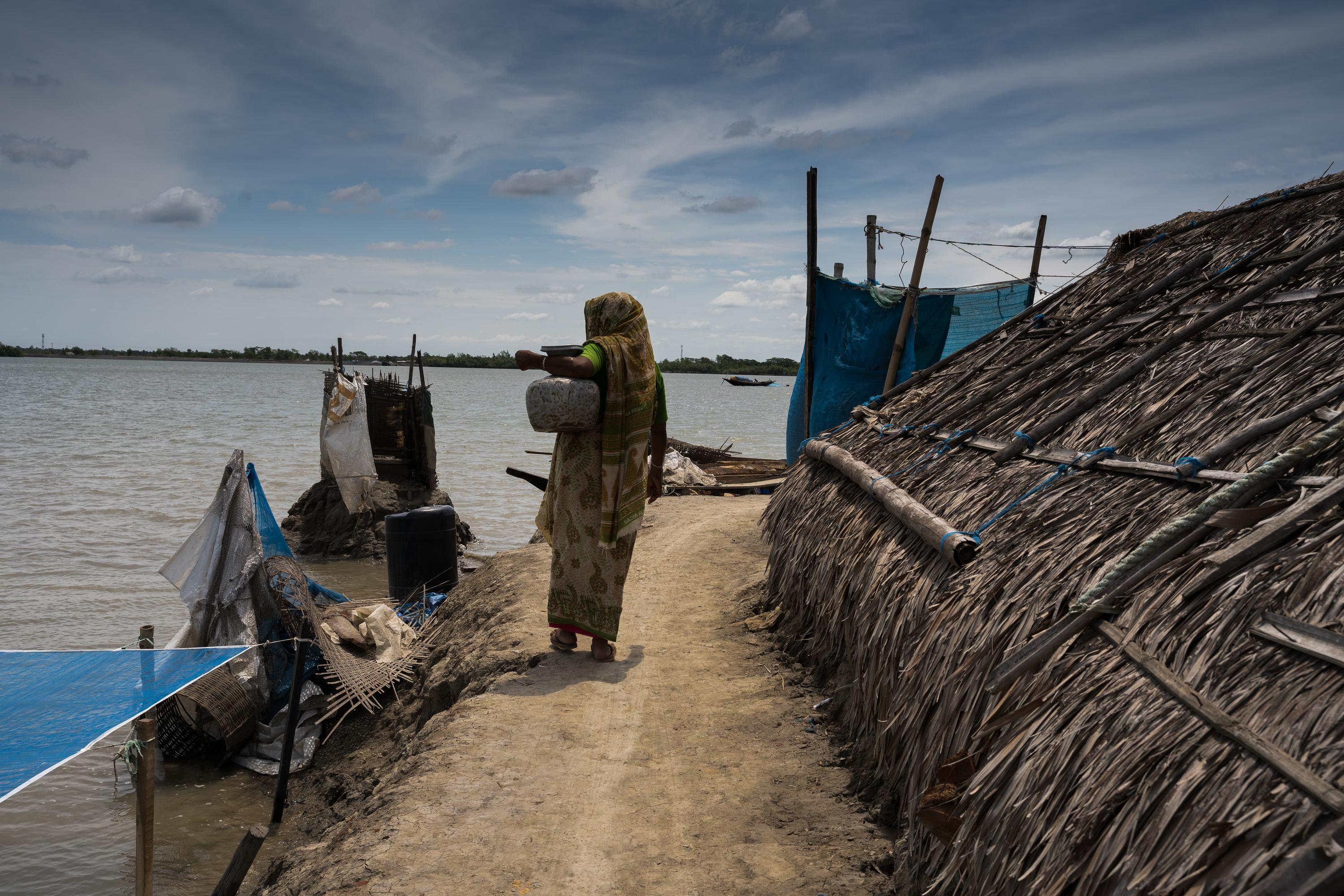 A woman carries fresh water along an embankment, while saline water swells in the background.