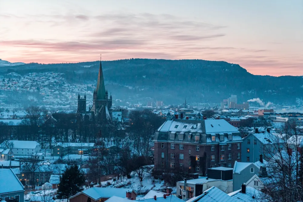 A winter view of Trondheim at sunrise, with snow-covered houses and the Nidarosdomen cathedral rising prominently in the centre.