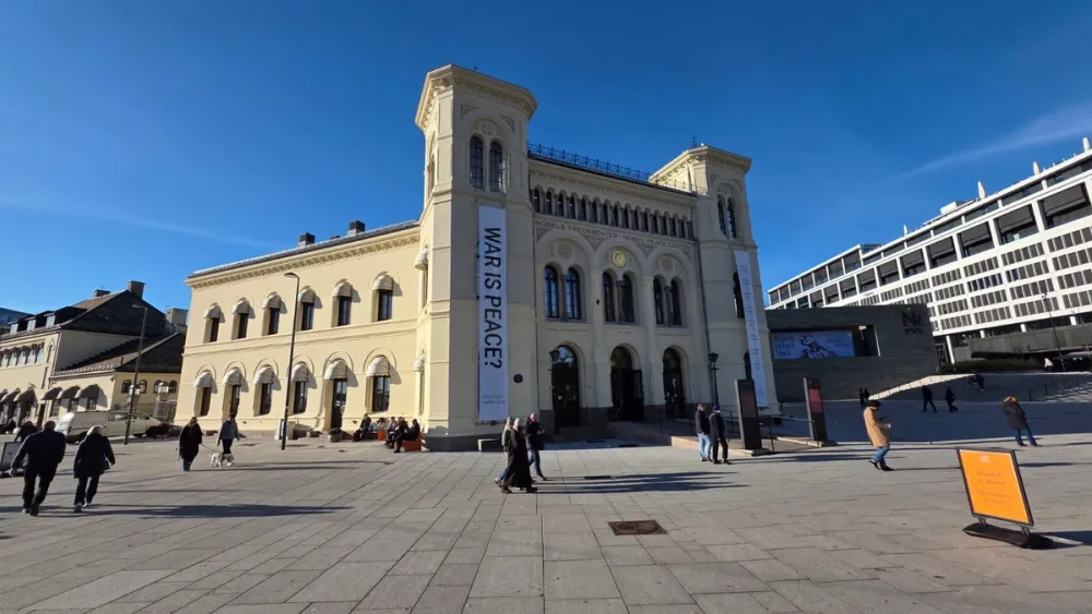 The Nobel Peace Center in Oslo, a cream-colored historic building under a clear blue sky. People walk across the square outside the museum, which houses exhibitions about peace, conflict resolution, and Nobel Peace Prize laureates.