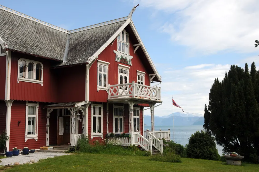 Red dragon style villa in Balestrand, Norway, with white carved wooden details and a view of the fjord in the background.