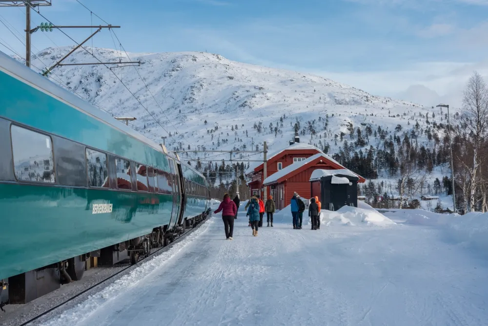 Voss train station in Western Norway in winter, with passengers walking beside the Bergen Line train and snow-covered mountains in the background.