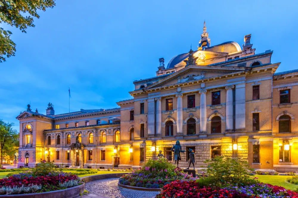 Evening view of the National Theatre in Oslo, with its illuminated neo-renaissance façade and surrounding flower beds.