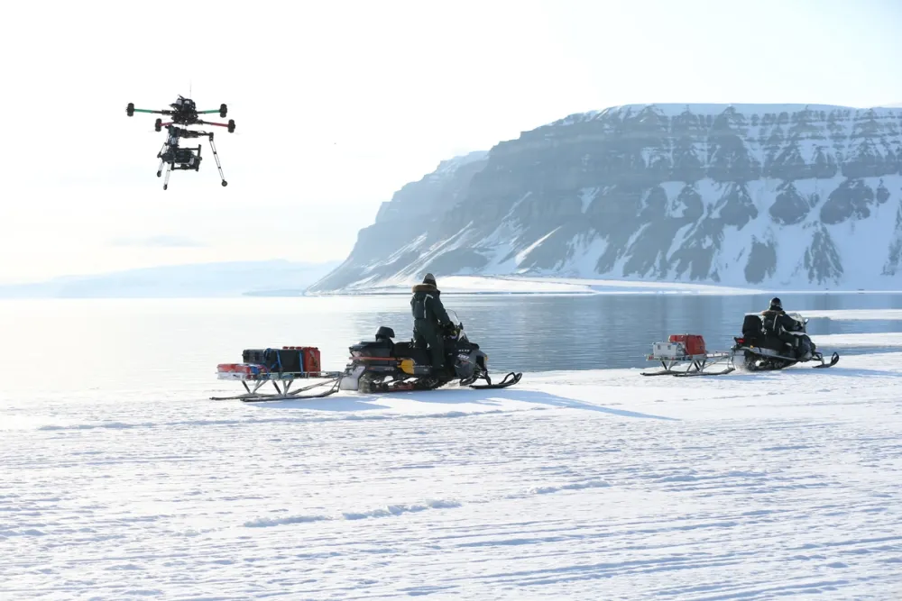 PolarX crew captures aerial footage using a heavy-lift drone near Svalbard’s icy coastline. With snowmobiles transporting equipment beside icy fjords and rugged cliffs, this scene highlights advanced Arctic cinematography techniques and mobile logistics in extreme filming environments.
