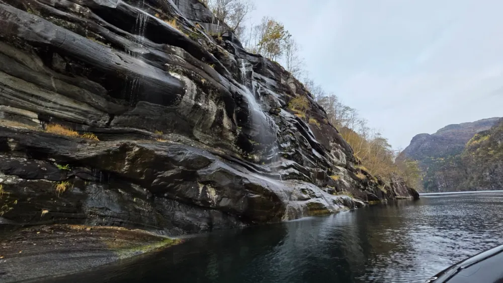 A view from the RIB boat in Hardangerfjord, showing dark rock cliffs with thin waterfalls trickling down into calm, reflective water. The autumn landscape and rugged mountains in the distance capture the raw beauty of Western Norway.