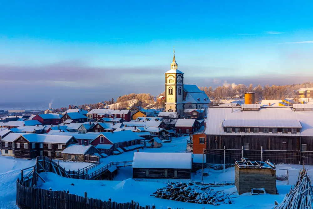 Winter view of the UNESCO World Heritage town of Røros, Norway, with colorful wooden houses and snow-covered rooftops under a clear blue sky.