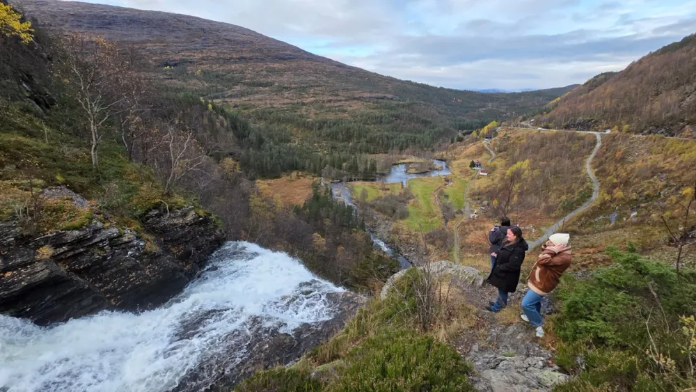 View from a cliffside in Hardanger showing a rushing waterfall cascading into the valley below. Participants from the fam trip stand at the edge, looking out over the river, winding road, and patchwork of forests and fields framed by mountains.