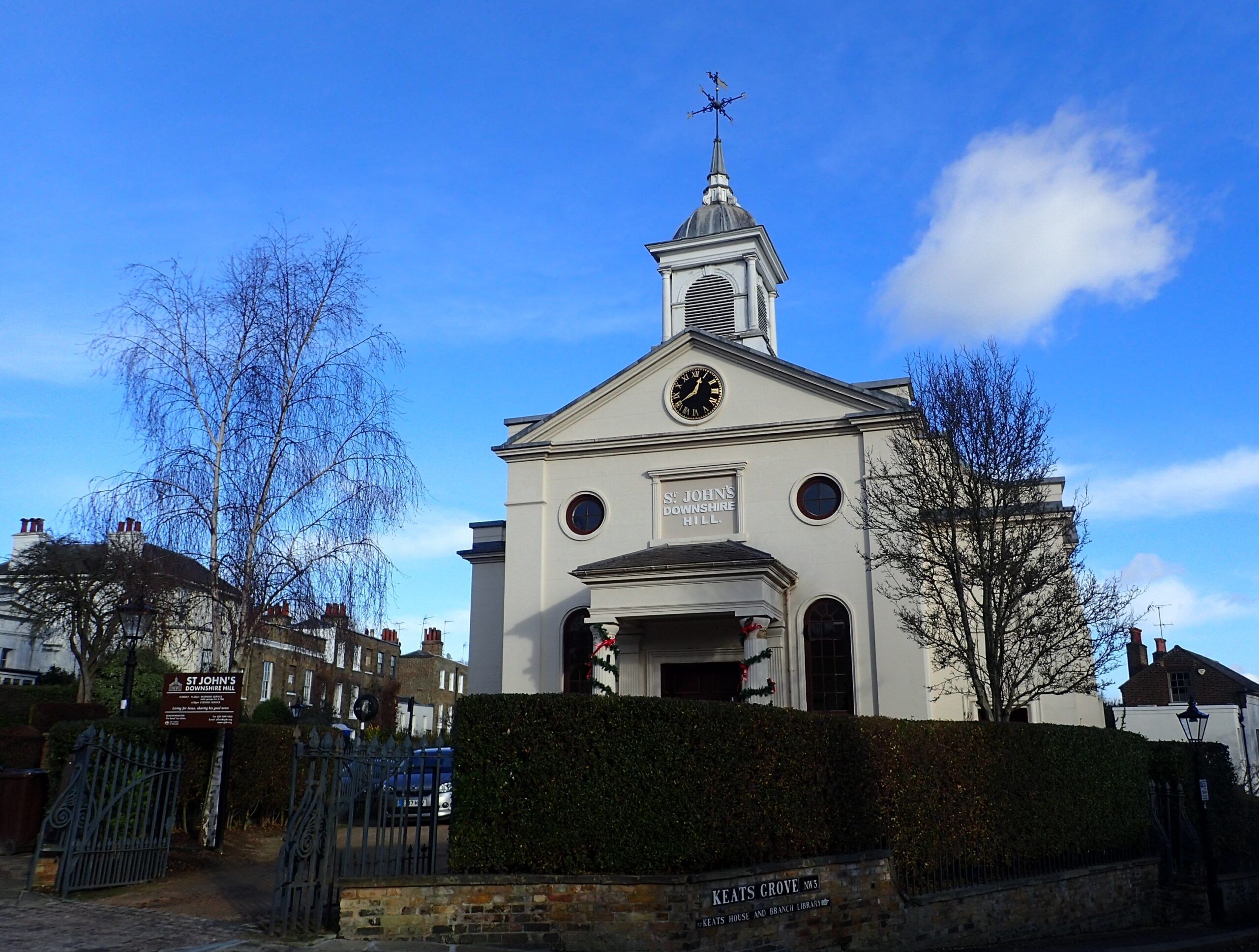 St John's Downshire Hill church with white facade, clock, steeple and cross, surrounded by bare trees and brick wall.